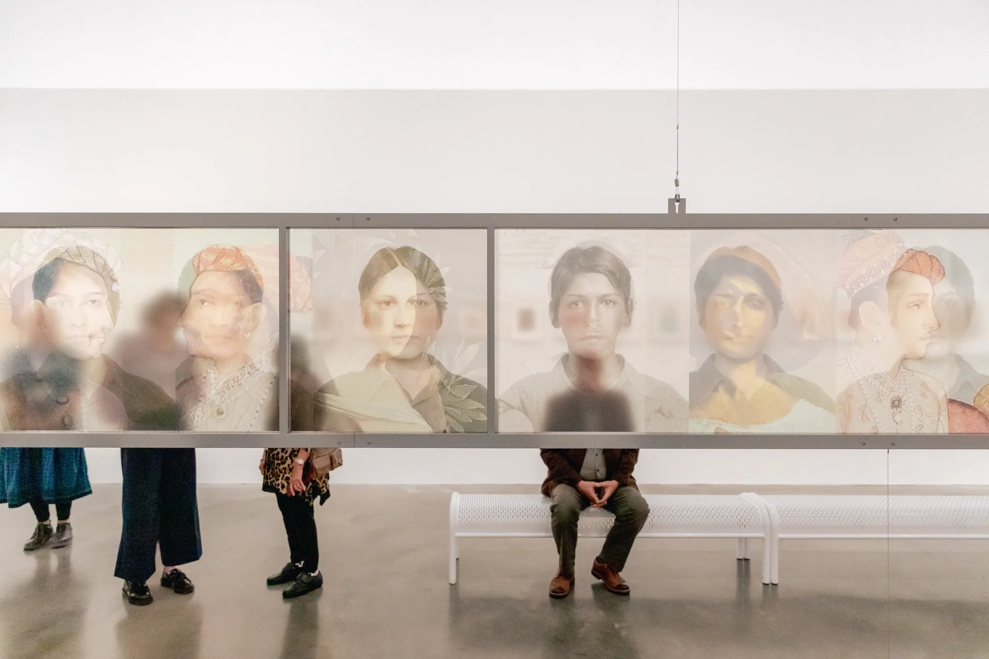 People observing artwork of multiple portraits behind glass, within an art gallery.