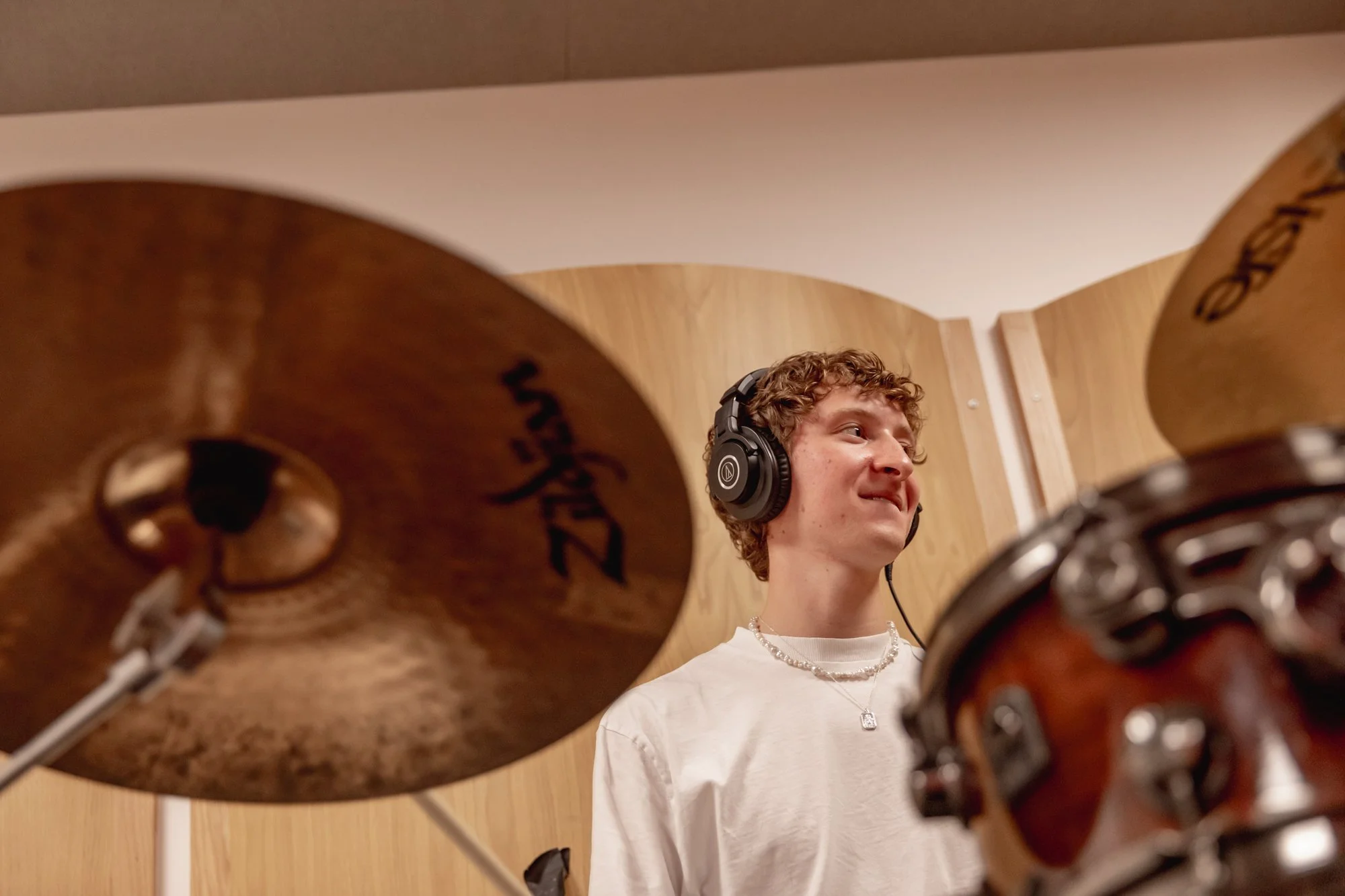 Young man with curly hair wearing headphones, sitting behind a drum set in a recording studio with wooden panels.