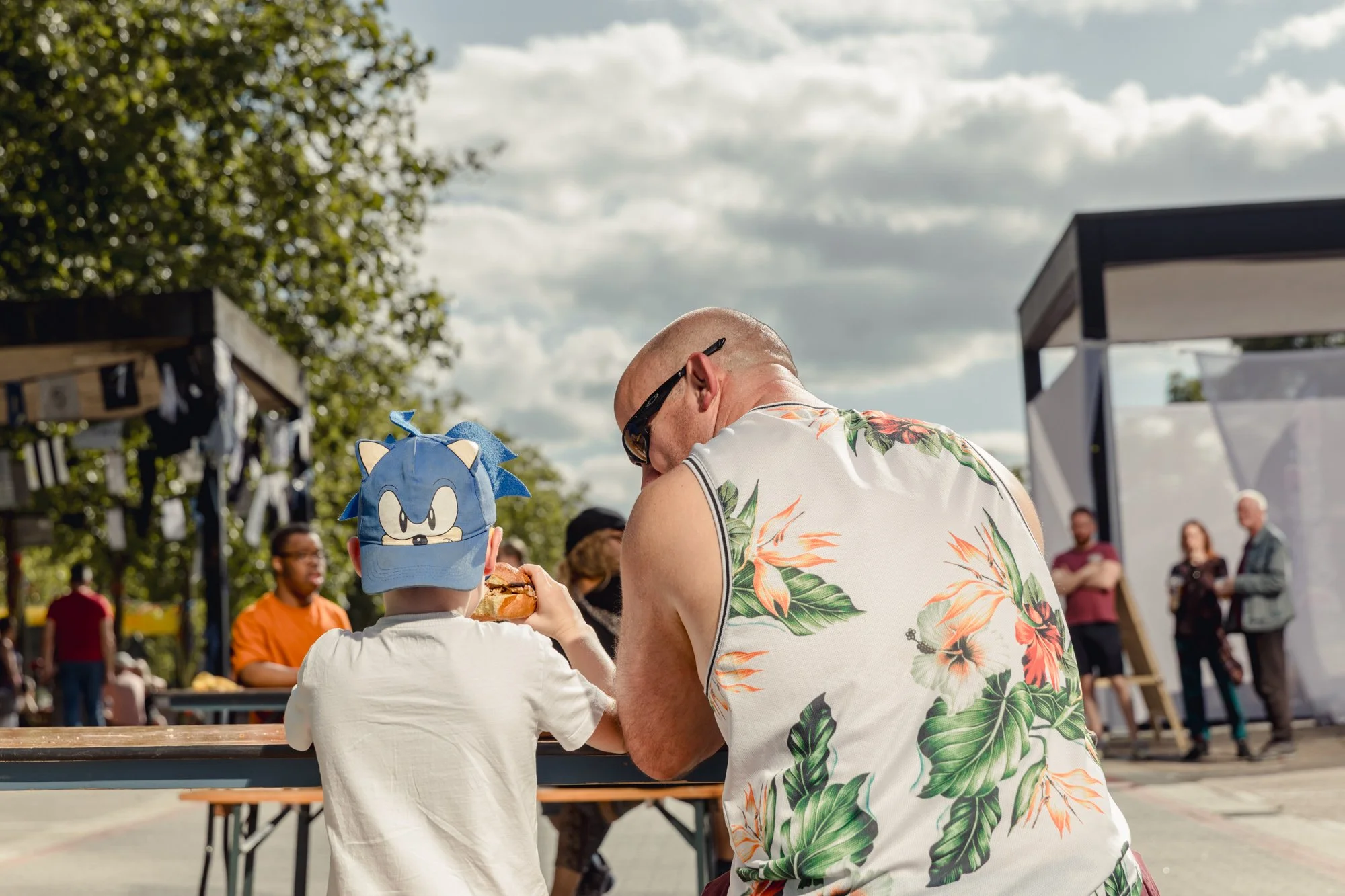 A man and a child sitting at a table outdoors during a sunny day. The child is wearing a Sonic the Hedgehog cap. In the background, other people are standing and talking, and there are trees and a structure with a glass or plastic enclosure.