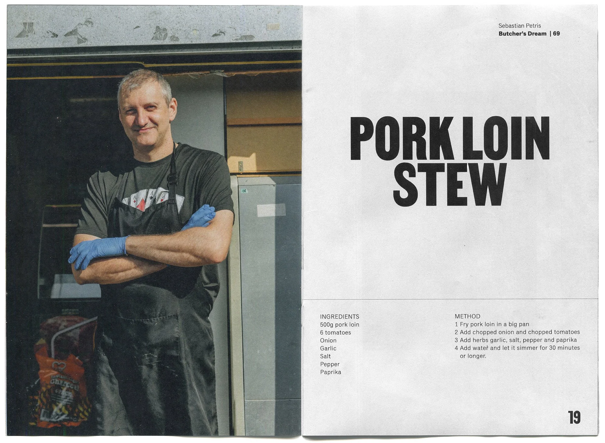 A man in a black apron and blue gloves standing with crossed arms in a kitchen or outdoor setting, smiling at the camera, with a stove and a bag of chips in the background.