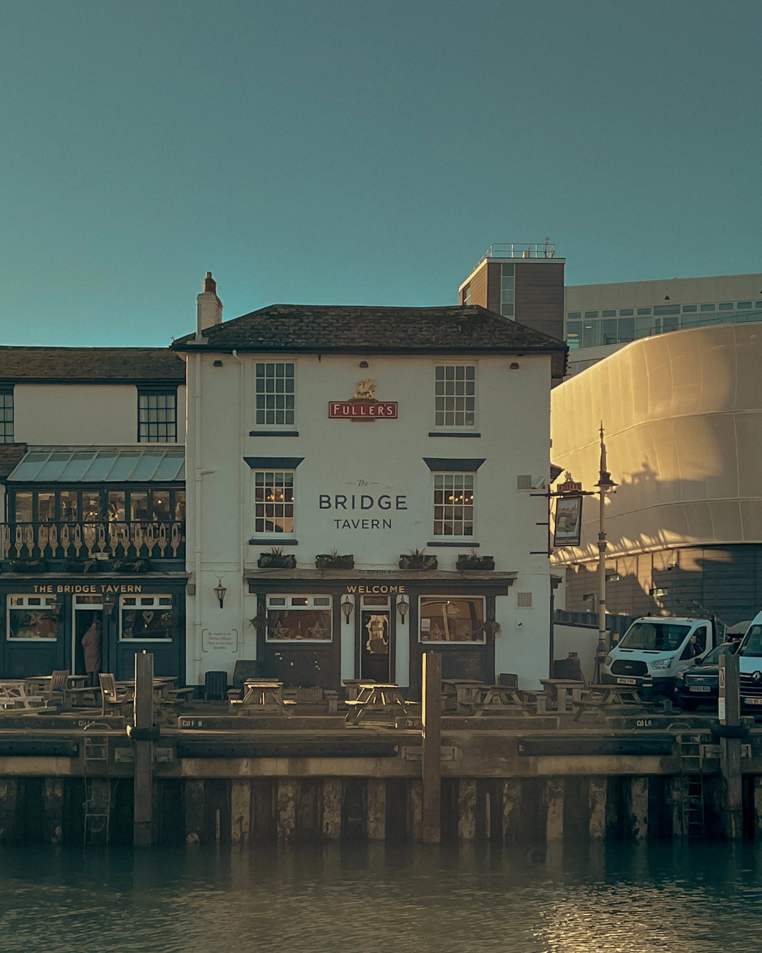 Front view of a white building named 'The Bridge Tavern' situated by water, with outdoor seating and a sign welcoming visitors, with city buildings in the background.