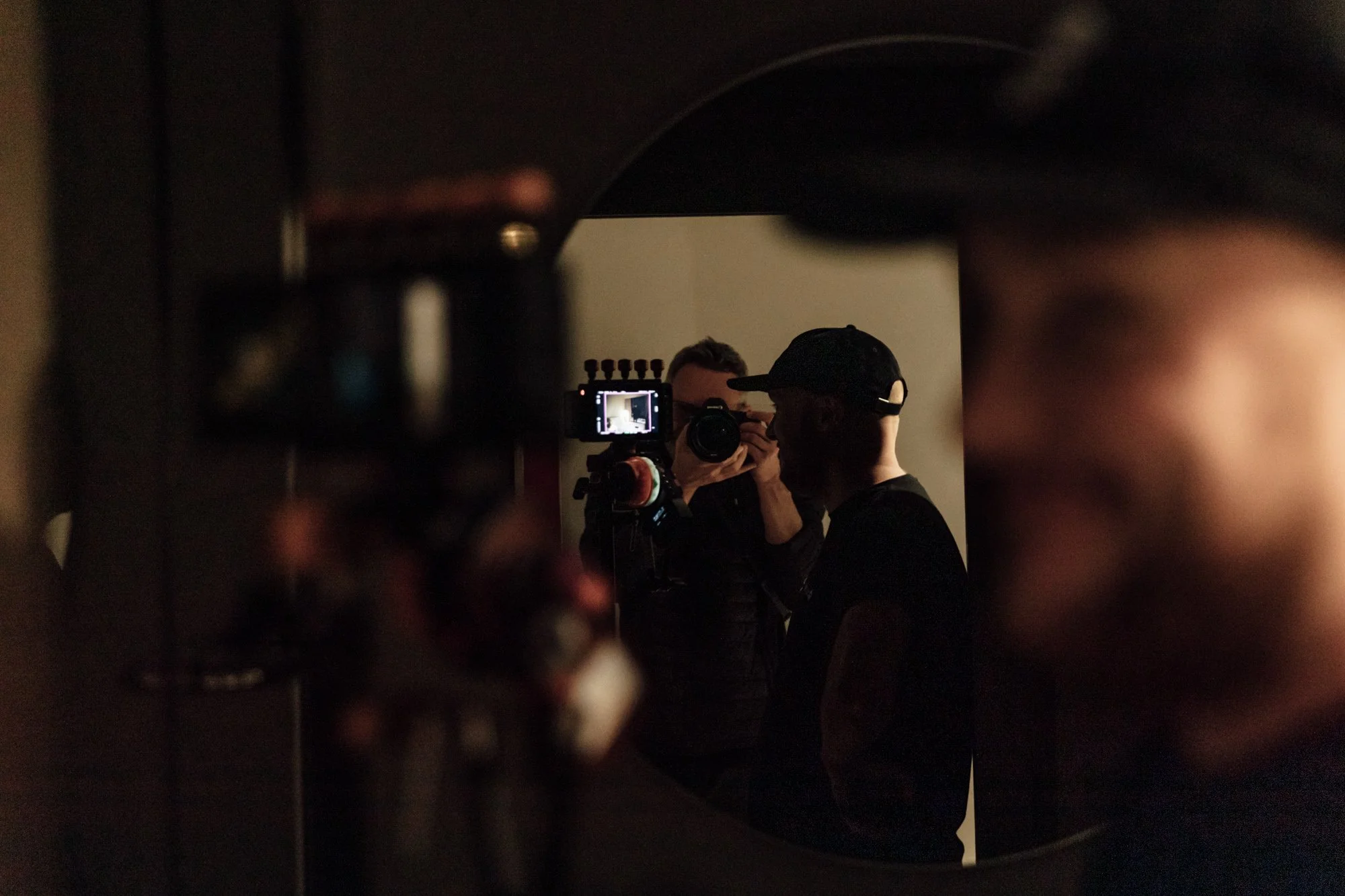Two individuals, one holding a camera, are reflected in a circular mirror. The scene appears to be in a dimly lit room.