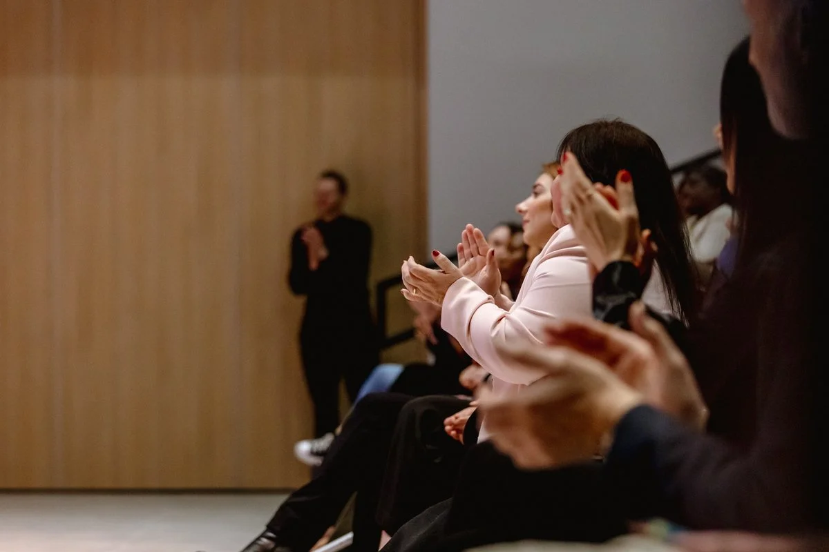 Women sitting and clapping in an audience at an indoor event.