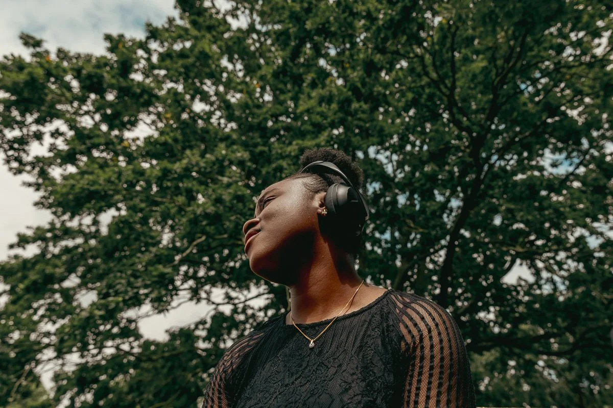 Young woman outdoors wearing large headphones, looking up with her eyes closed, against a backdrop of green trees.