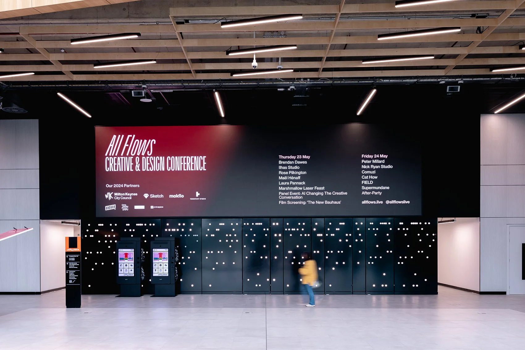 Large digital conference schedule screen for 'All Flows Creative & Design Conference' in a modern building lobby with a patterned black wall and minimalistic decor. A person in a yellow jacket is walking past.