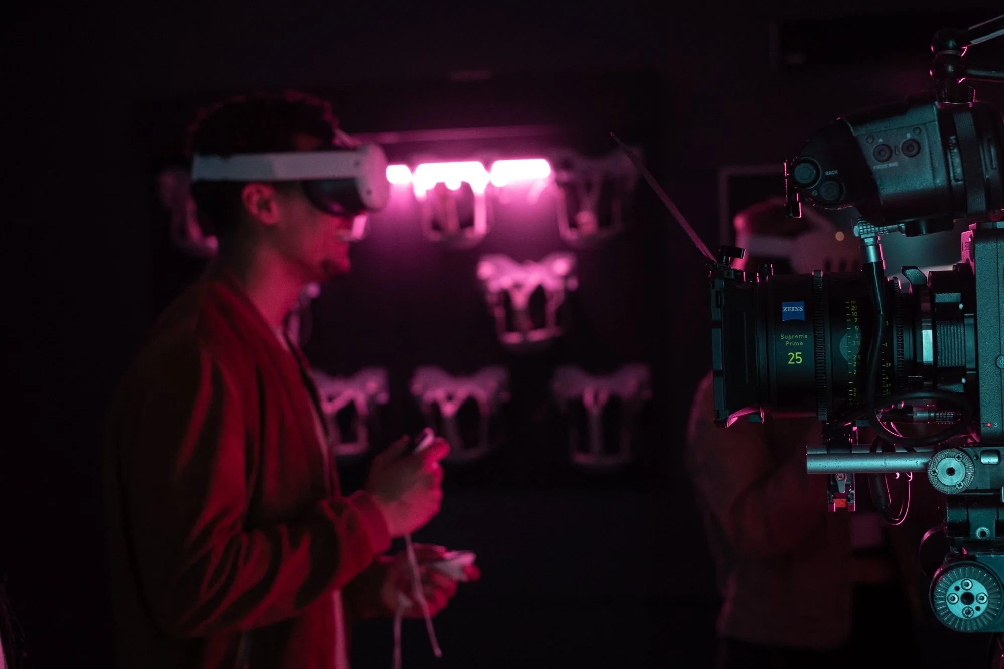 A person wearing a virtual reality headset and red jacket using a smartphone, with a professional video camera in the foreground and Nike shoes displayed on a black background with neon pink lighting.