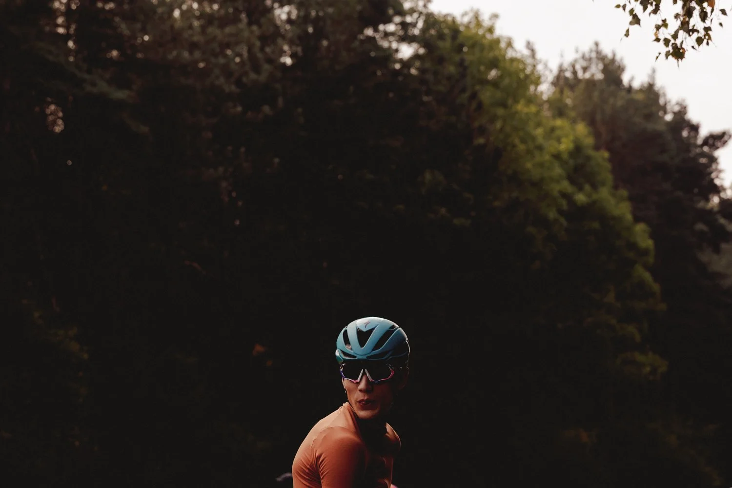 A woman wearing a blue bike helmet and sunglasses is outdoors surrounded by trees at dusk.
