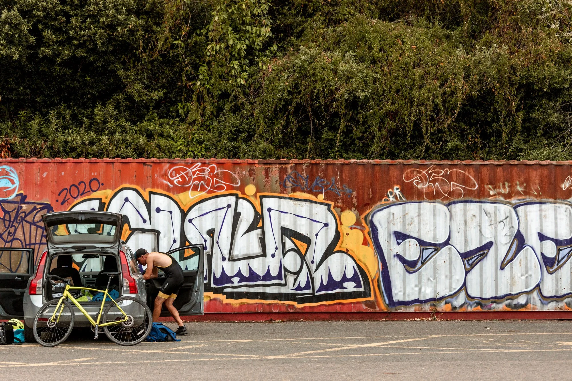 A man in black athletic clothing is packing a gray car with its trunk open, parked on an asphalt road. Next to the car is a yellow bicycle and some gear on the ground. Behind them is a red container with large white and purple graffiti, with green tr
