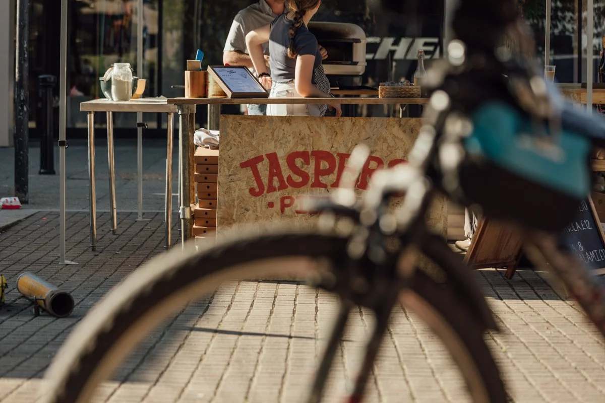 A food stand with a sign that reads "Jaspas" in front of a person preparing food on a grill, with a bicycle in the foreground and a woman with a kid at the stand.