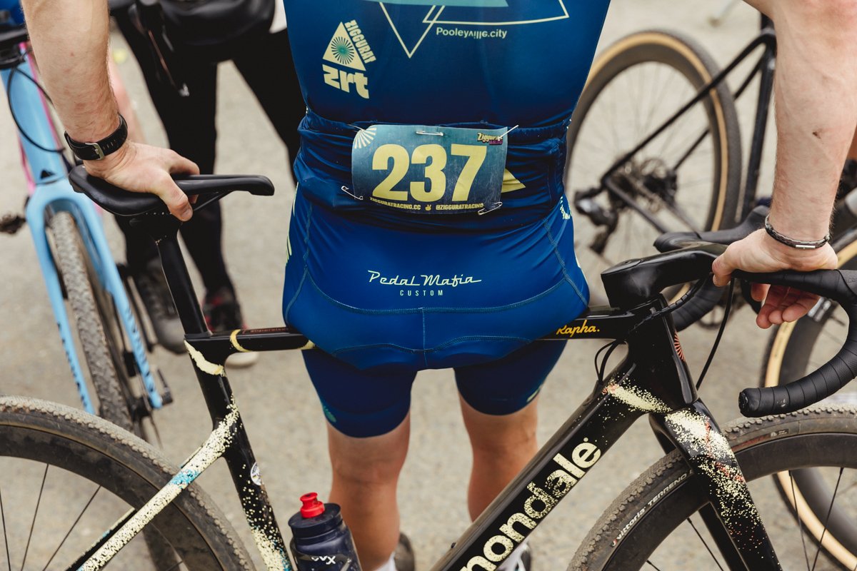 A cyclist standing with a black and gold Cannondale bike, wearing a blue cycling uniform with a race number 237 attached to the waist, and other bicycles in the background.