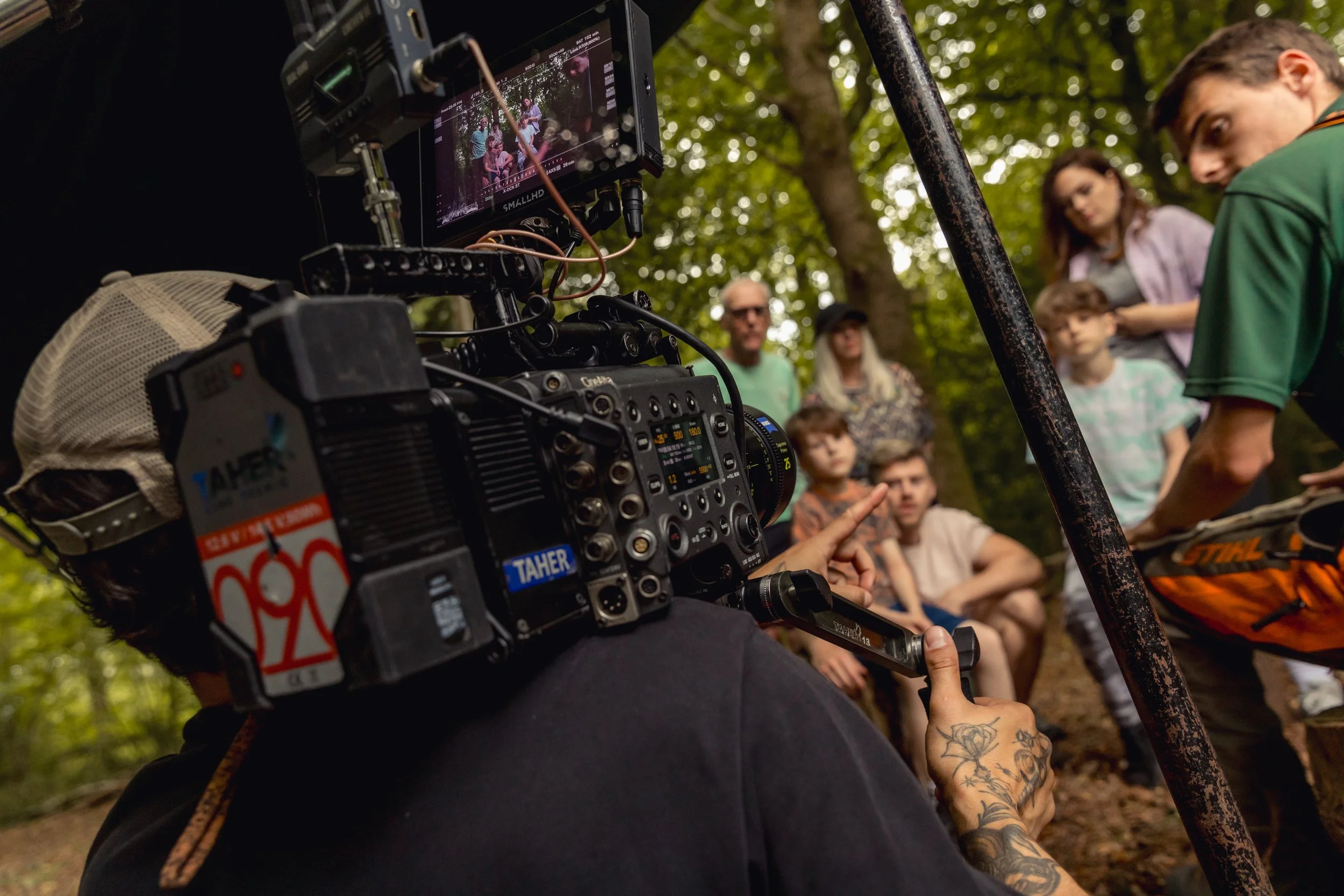 A film crew filming a scene outdoors in a wooded area with a vintage movie camera, while several actors or participants sit and stand in the background.