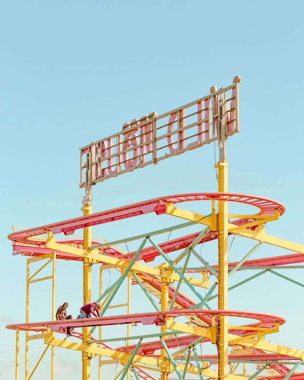 A vintage roller coaster with pink tracks and yellow supports, featuring a large sign at the top that says 'CUPID'. Two children are climbing on the coaster's track amidst a clear blue sky.