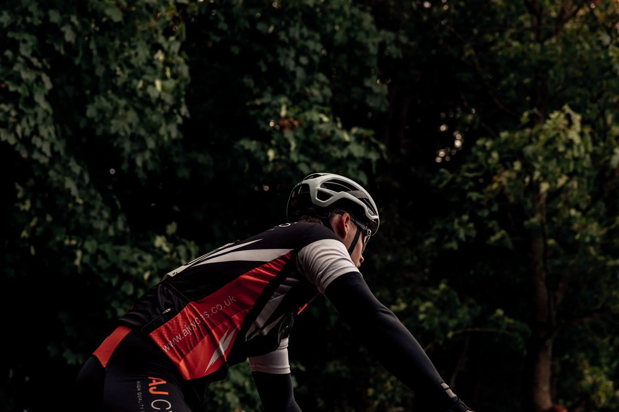 A cyclist wearing a black, white, and red cycling jersey and a white helmet riding outdoors against a background of green trees.