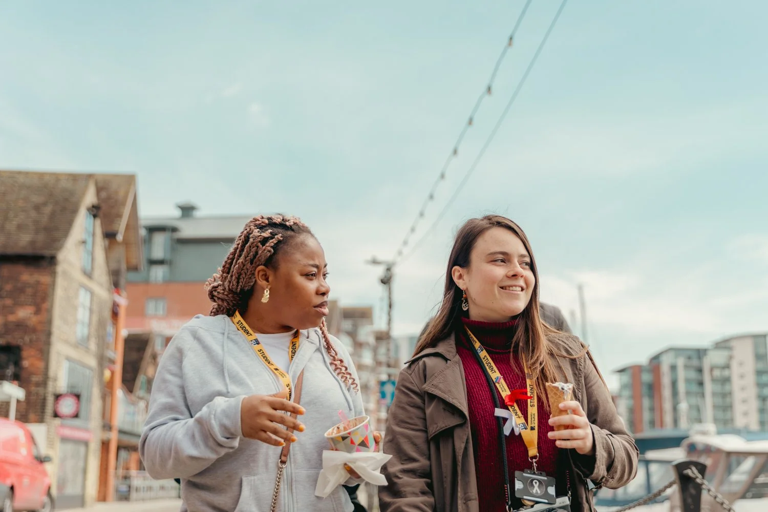 Two women walking outdoors in a city, holding ice cream cones, wearing lanyards with badges, with buildings and power lines in the background.