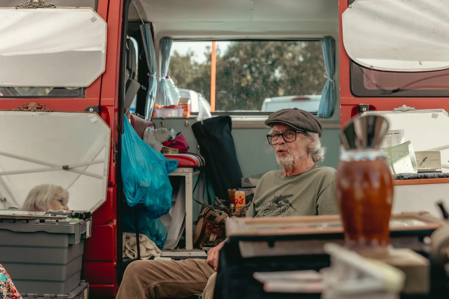 An elderly man with glasses and a cap sitting inside a red camper van, with a cluttered interior including bags, boxes, and kitchen items.