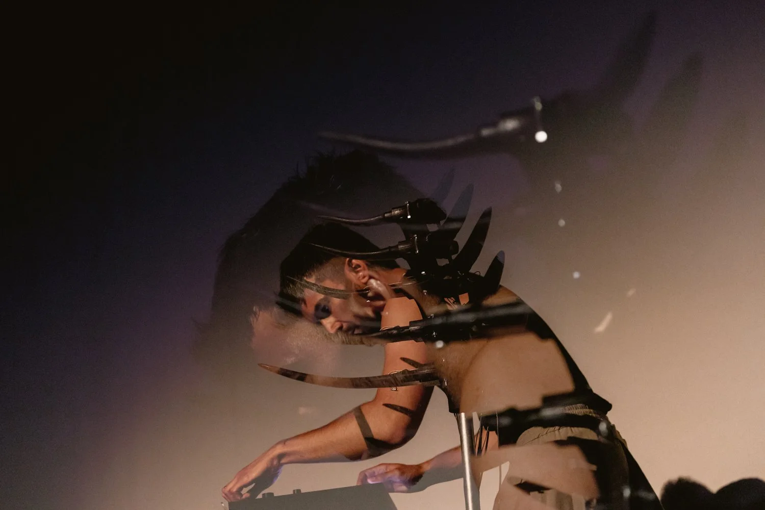 A DJ with tattoos and glasses is performing on stage with a dark background, leaning over a turntable.