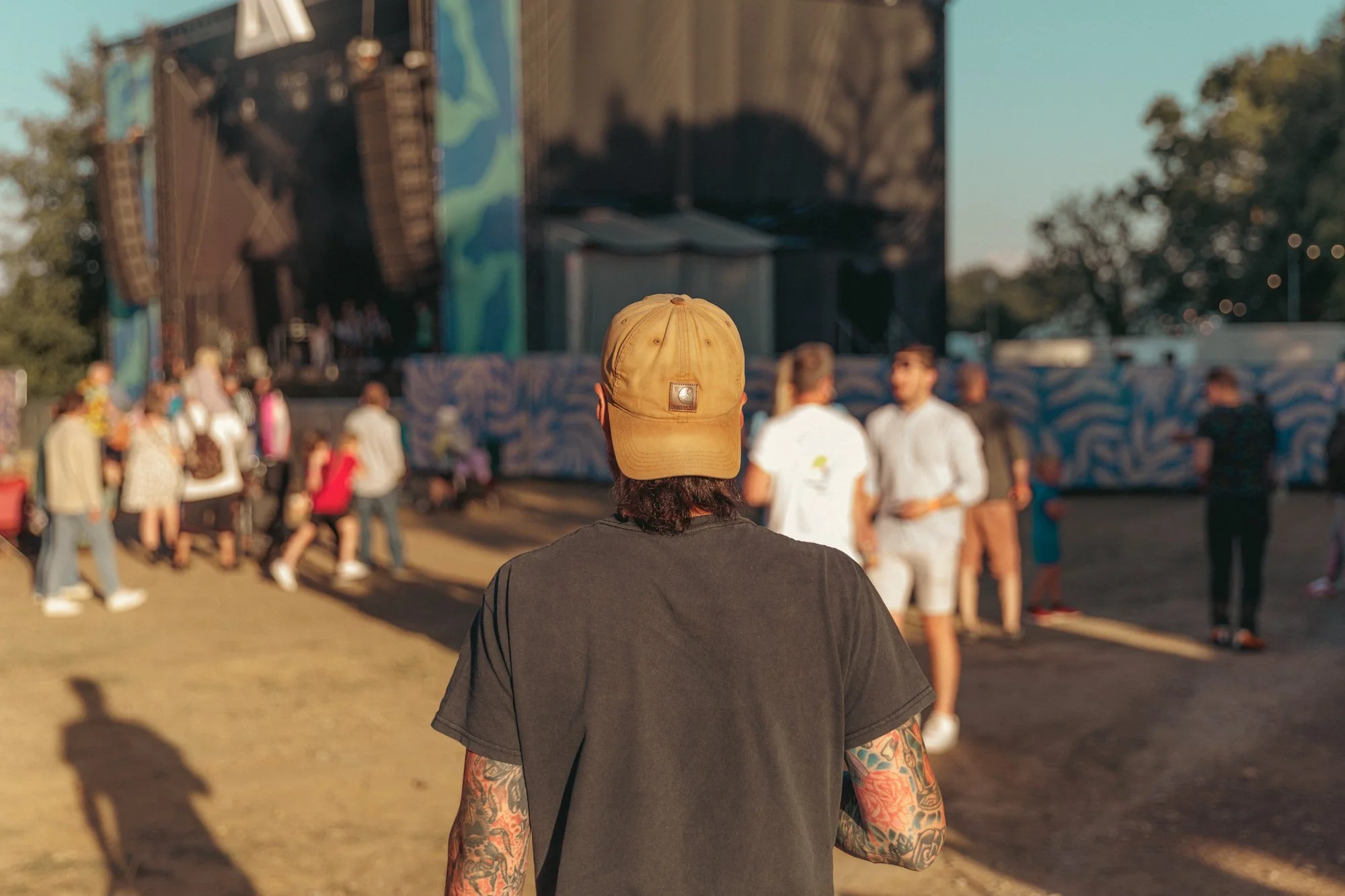 Person wearing a tan cap and black t-shirt with tattoos on arms, seen from behind, at an outdoor concert with a stage and audience in the background.
