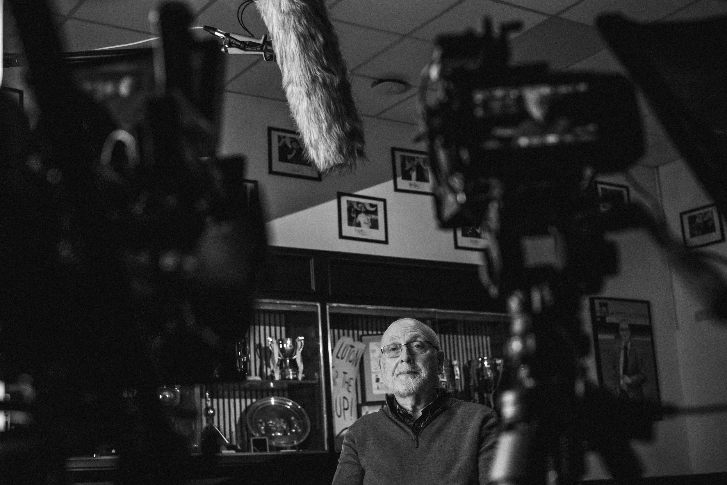 Black and white photo of an older man with glasses and a beard, sitting in a room surrounded by cameras and recording equipment. The background has framed pictures on the wall and shelves with trophies and decorative items.