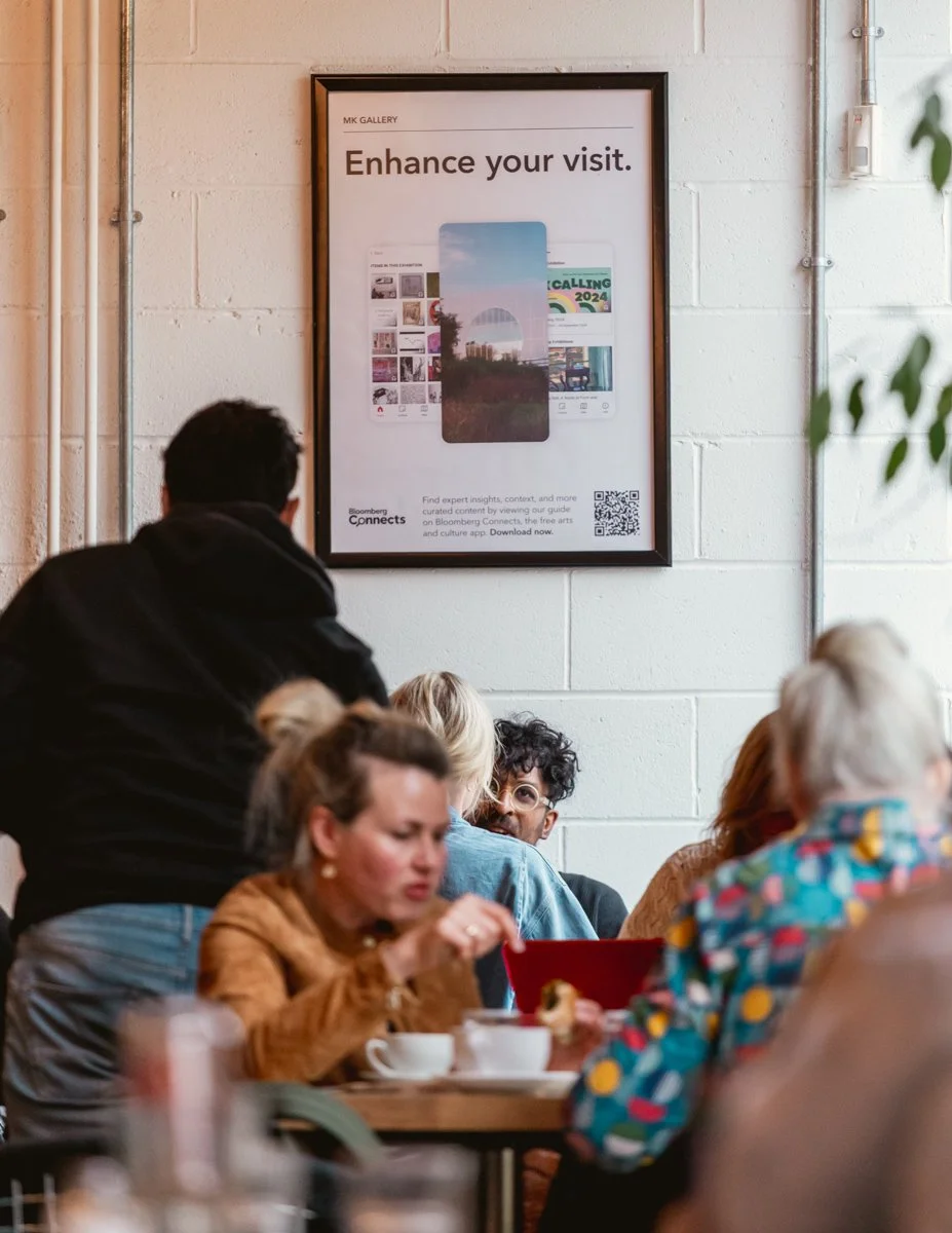 People sitting at tables in a cafe or restaurant with a framed poster on the wall that reads 'Enhance your visit.'.