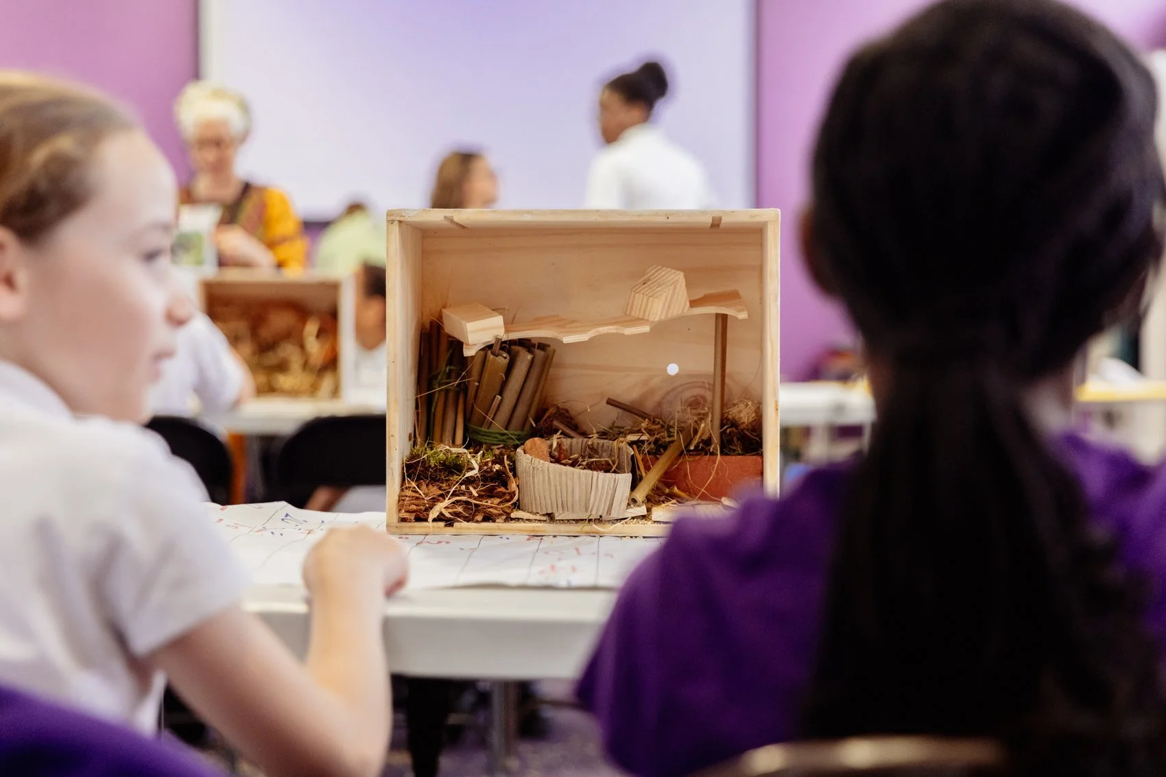 Children observing a small diorama of a natural scene with plants, rocks, and miniature structures at a classroom or exhibit setting.