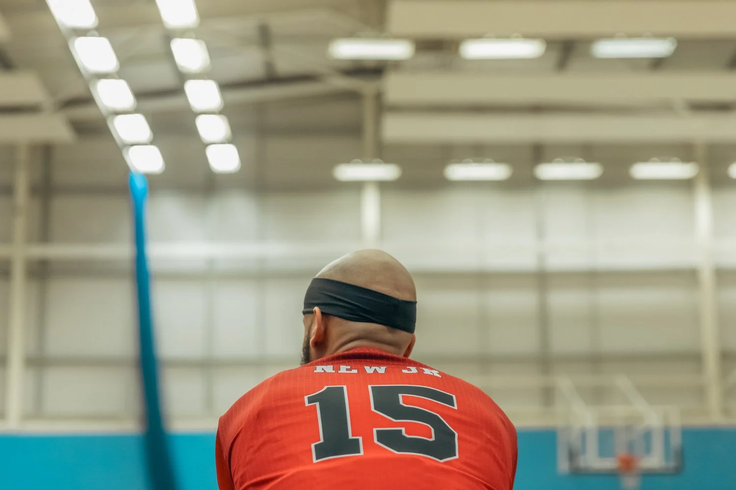 Back of a basketball player wearing a red jersey with the number 15 and a black headband in an indoor gymnasium.