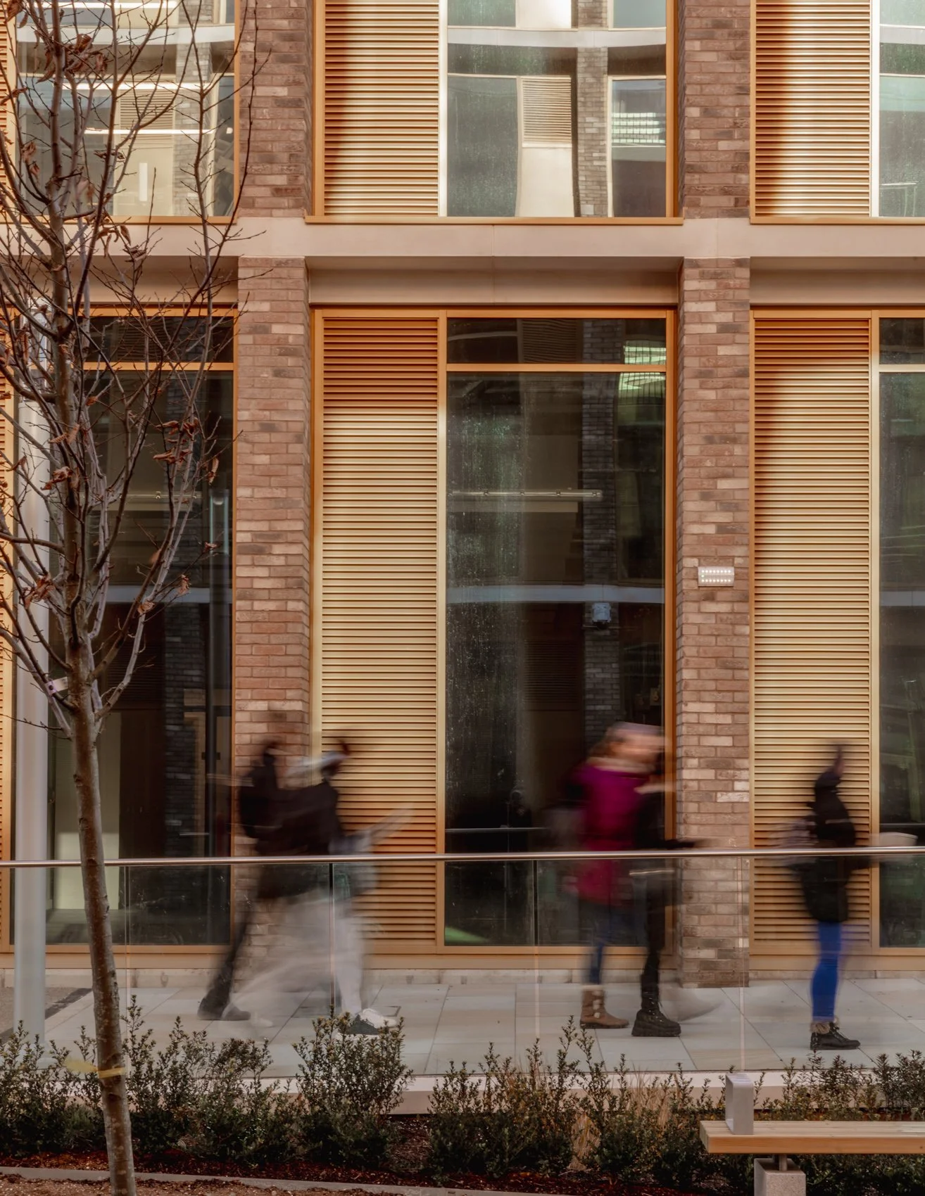 Blurred people walking in front of a modern building with large windows and wooden external shutters, a tree on the left and small bushes at the bottom.