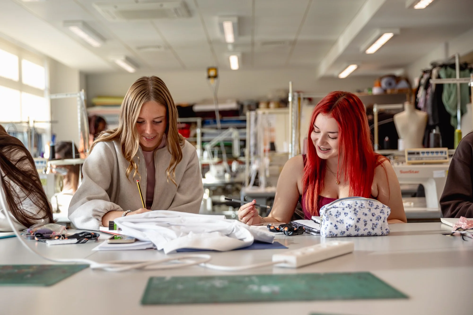Two women sit at a sewing table, smiling and working on fabric and notebooks in a sewing workshop.