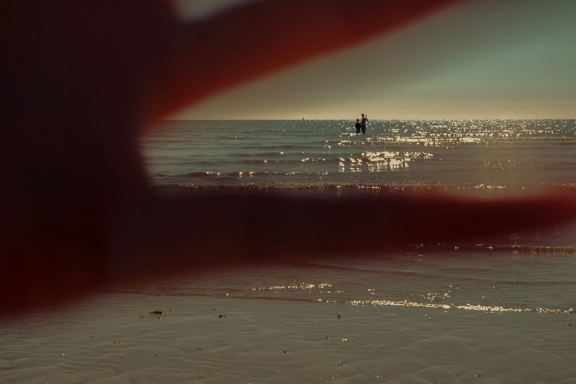 View of a couple standing in the ocean water near the shore, framed by a finger, at sunset with sunlight reflecting on the water.