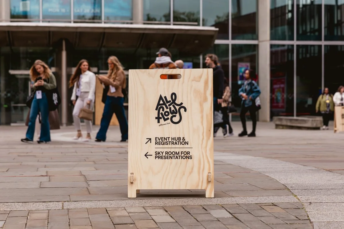 A wooden signboard with event information, placed on a paved outdoor area near a modern glass building, with people walking in the background.