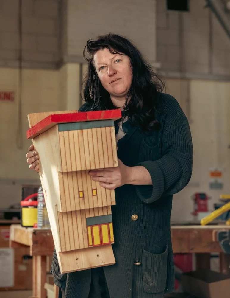 Woman with black hair holding a tall wooden house with painted details in a workshop.