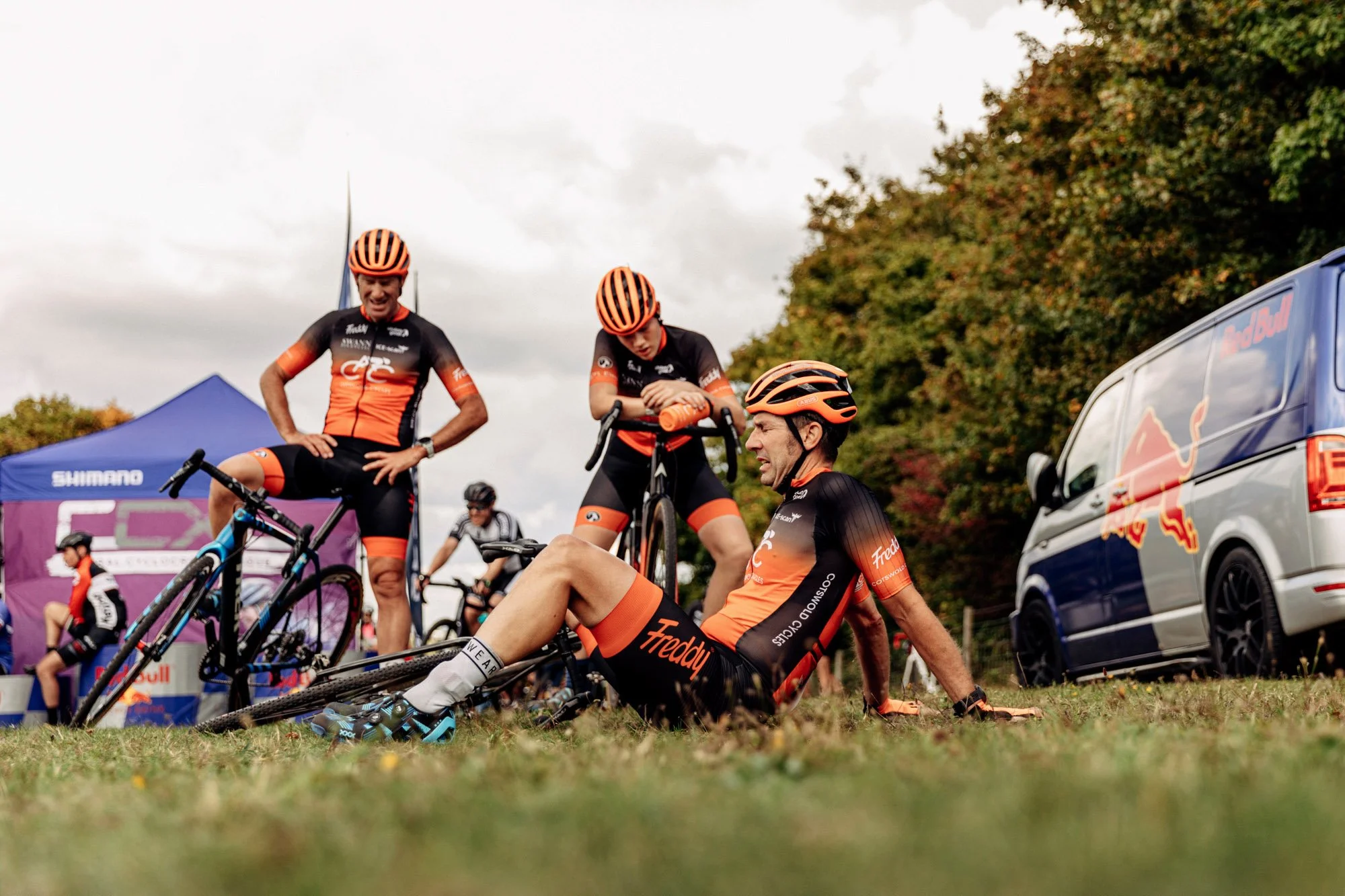 Three cyclists wearing orange and black jerseys and helmets taking a break at a cycling event. One is sitting on the ground, one is standing and leaning on a bicycle, and the third is standing with a bicycle. There is a Red Bull vehicle and event ten