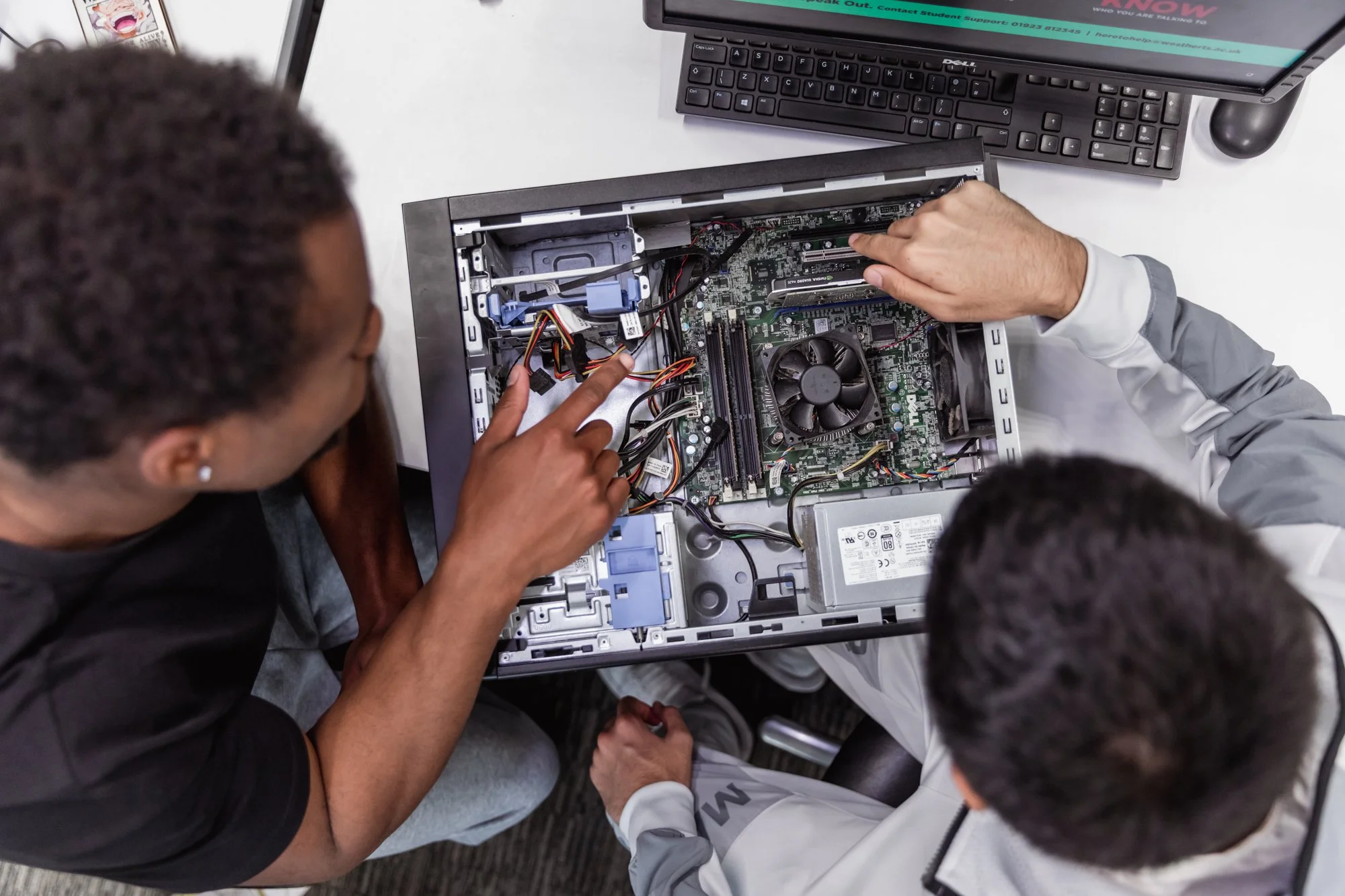 Two individuals working inside a desktop computer, with one pointing at the circuit board, at a desk with a monitor, keyboard, and mouse.