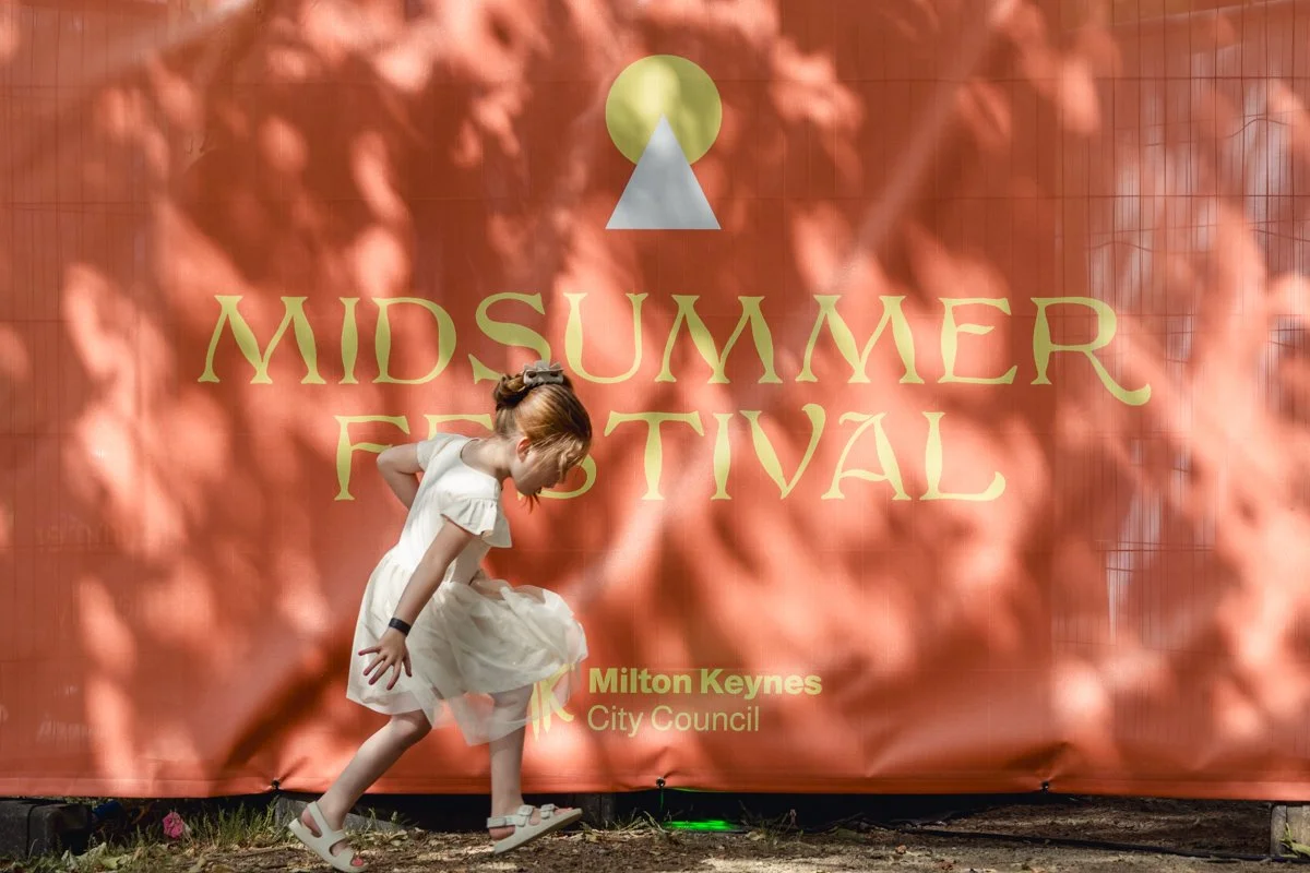 A young girl in a white dress and sandals walking in front of a large red banner that reads "Mid-Summer Festival" with a logo and the words "Milton Keynes City Council."