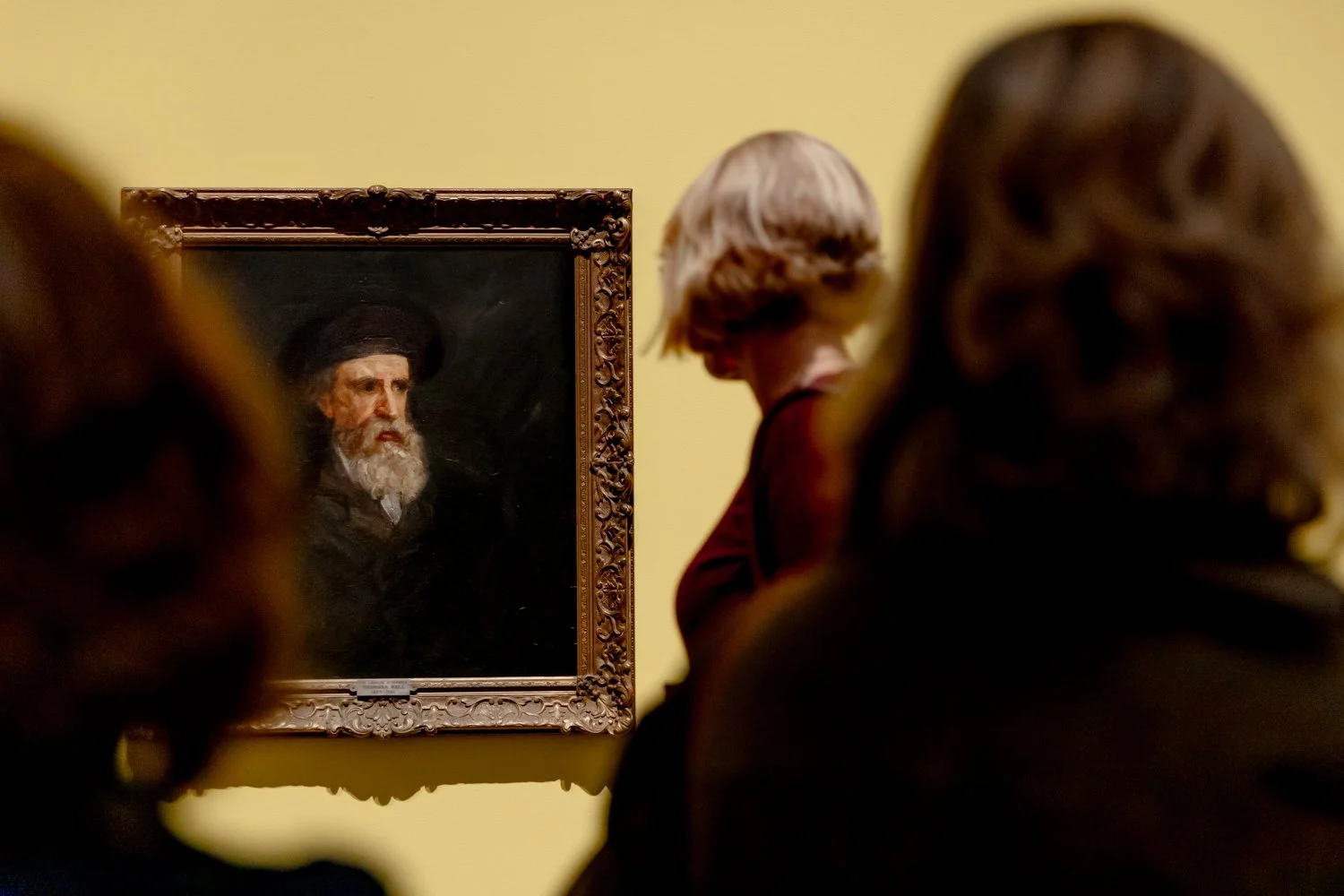 People viewing a portrait of an older man with a beard and hat in an art gallery.