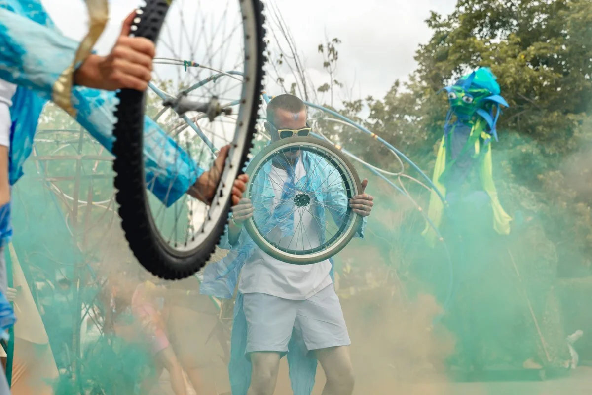 Group of people outdoors amid colorful smoke, holding bicycles and cycling gear, with trees in the background.