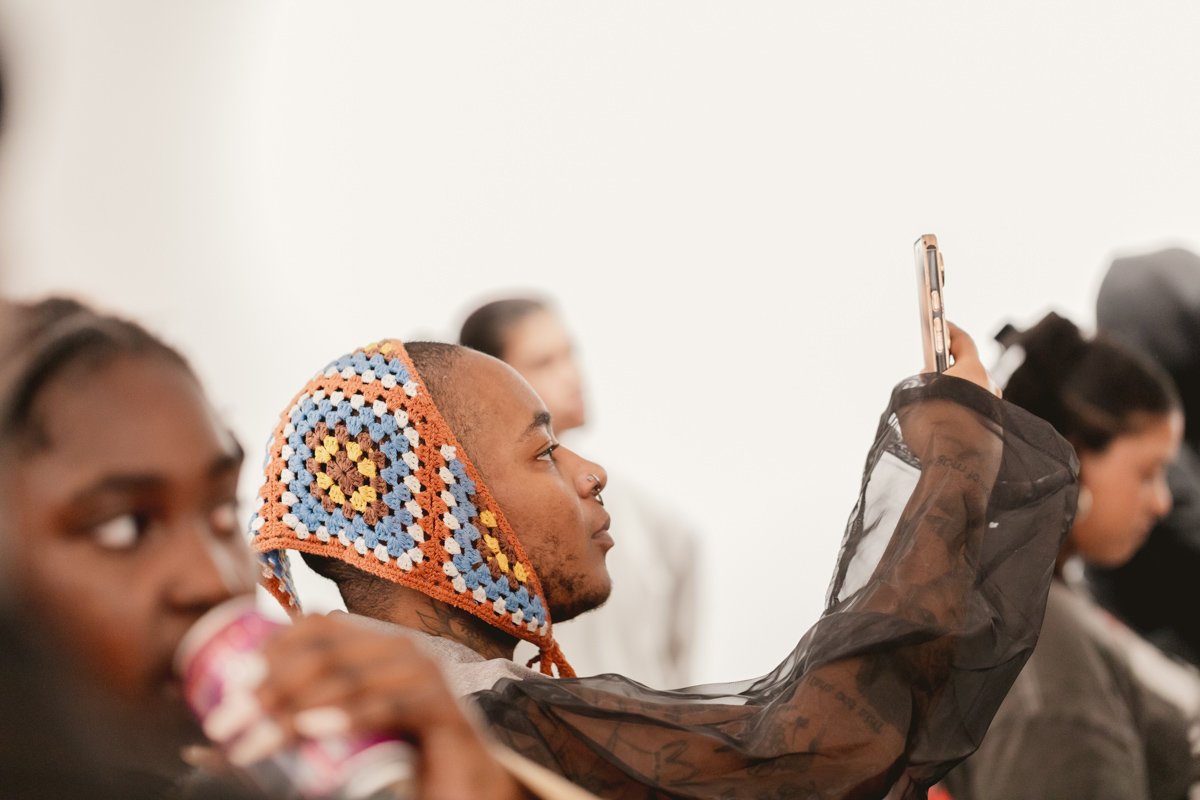 A young man with a nose ring wearing a colorful crochet hat and a sheer black covering is taking a photo with his phone. Other people are visible around him, including a young woman drinking from a cup in the foreground and others in the background.