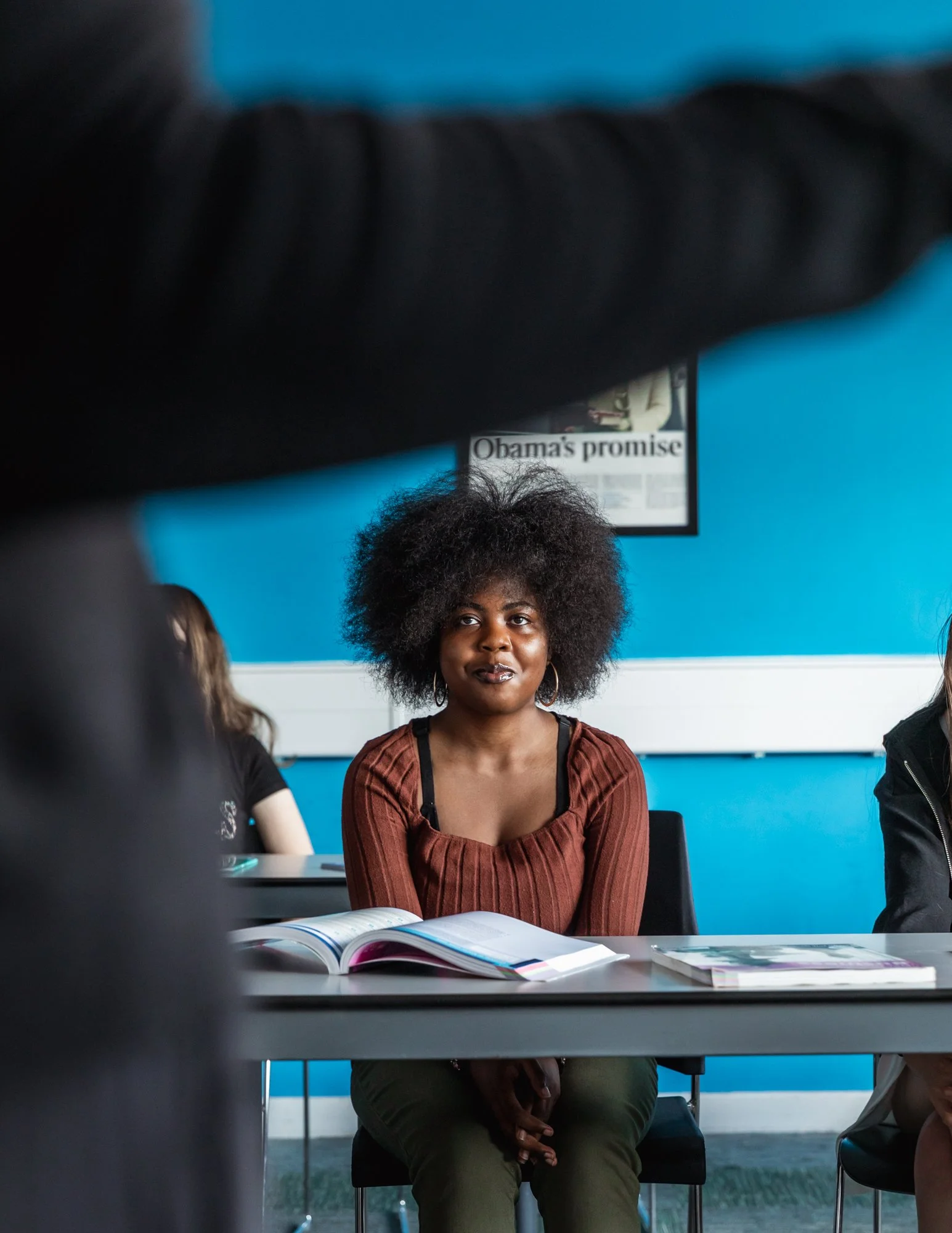 A woman with curly black hair wearing a rust-colored top sitting at a table with open books in front of her, in a room with blue walls and a sign that reads 'Obama's promise' in the background.