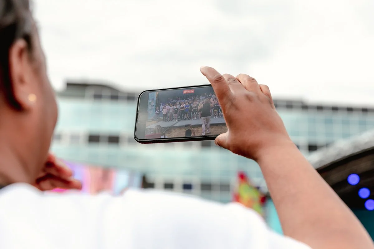 Person taking a photo or recording a video of a group of people on a stage outdoors with a smartphone.