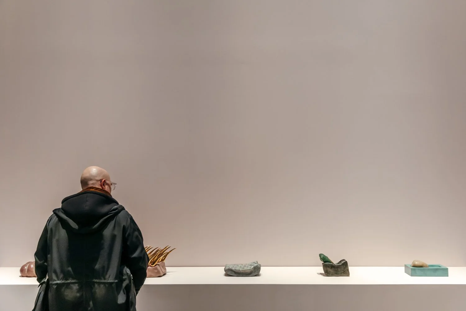 A man with a bald head and glasses, wearing a black jacket, stands in front of a white display shelf with various objects, including sculptures and decorative items, against a plain beige wall.