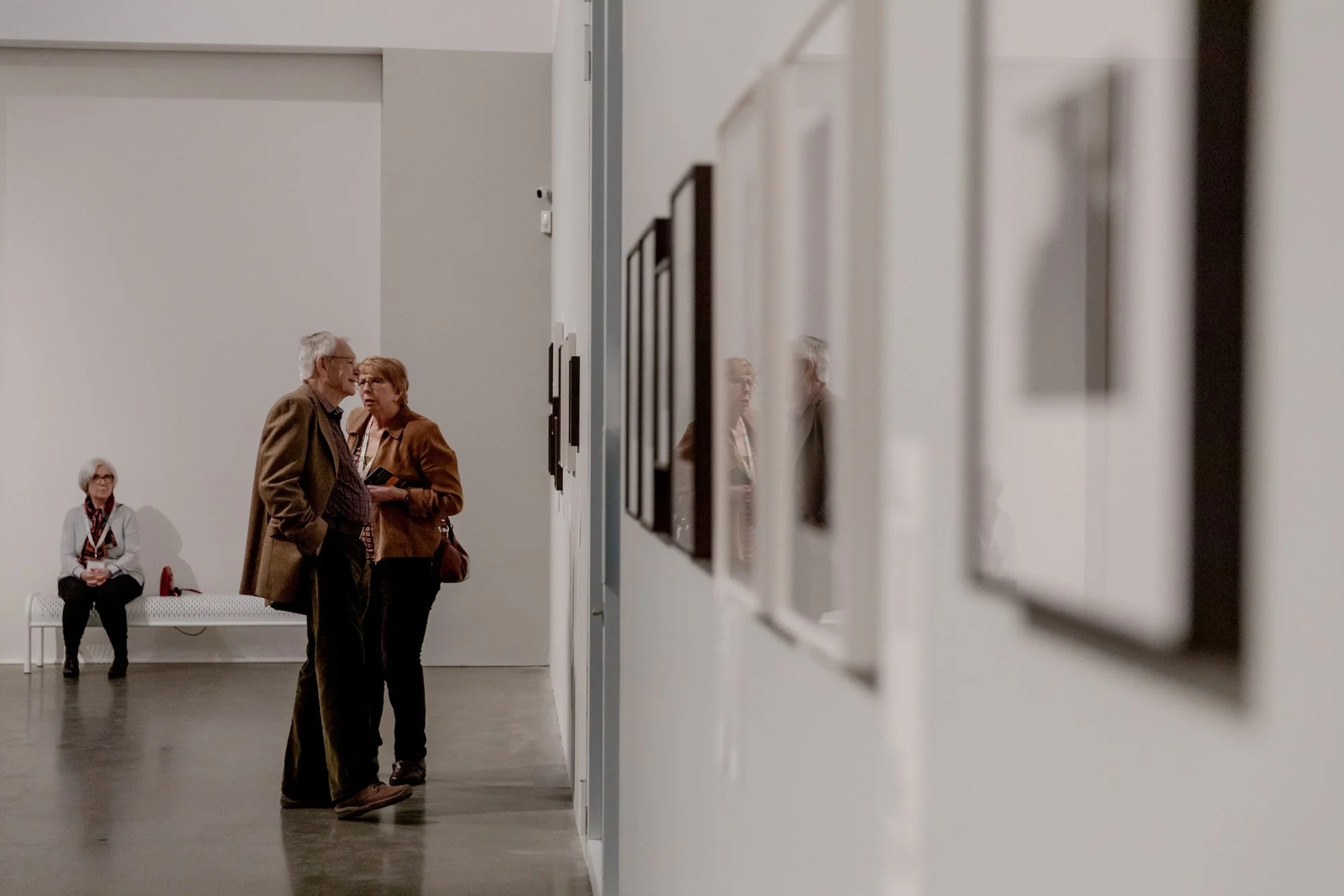 Two elderly individuals are engaged in conversation in an art gallery, with another person seated on a bench nearby. The gallery walls display framed artwork.