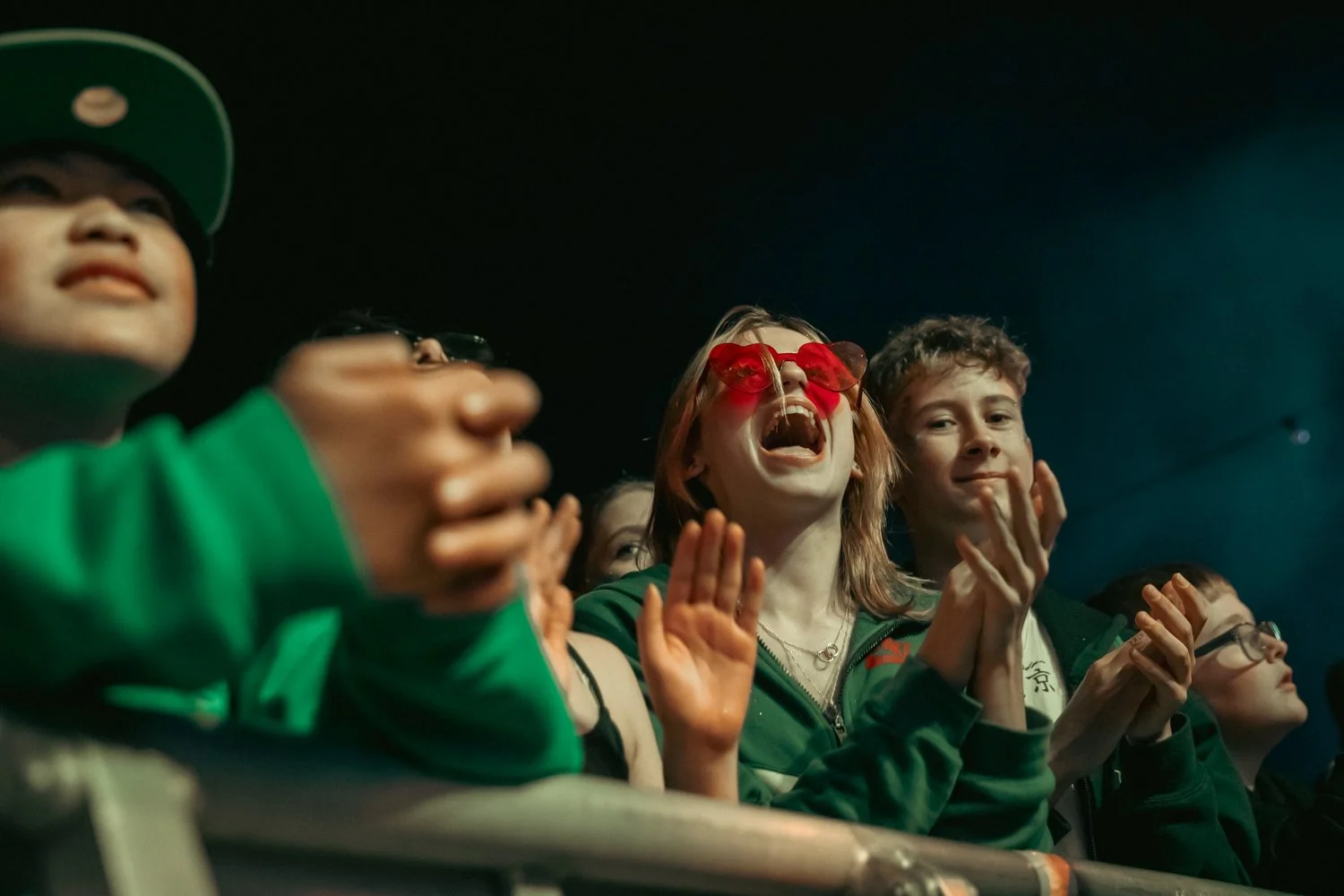 Group of children and teens at a concert or event, smiling, clapping, and cheering, with a dark background and some wearing sunglasses.