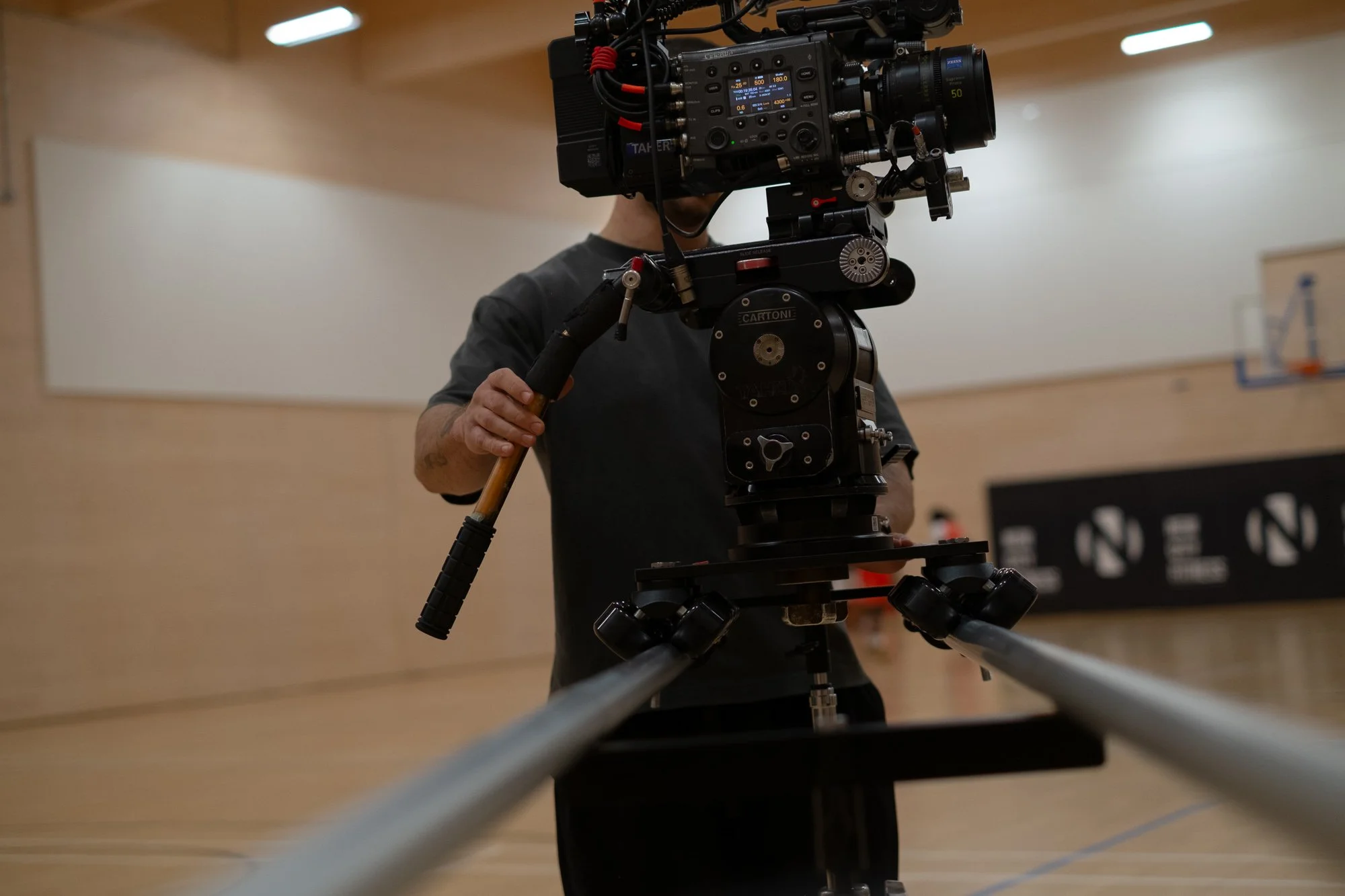 A person operating a camera mounted on a dolly track in an indoor gymnasium or sports hall.
