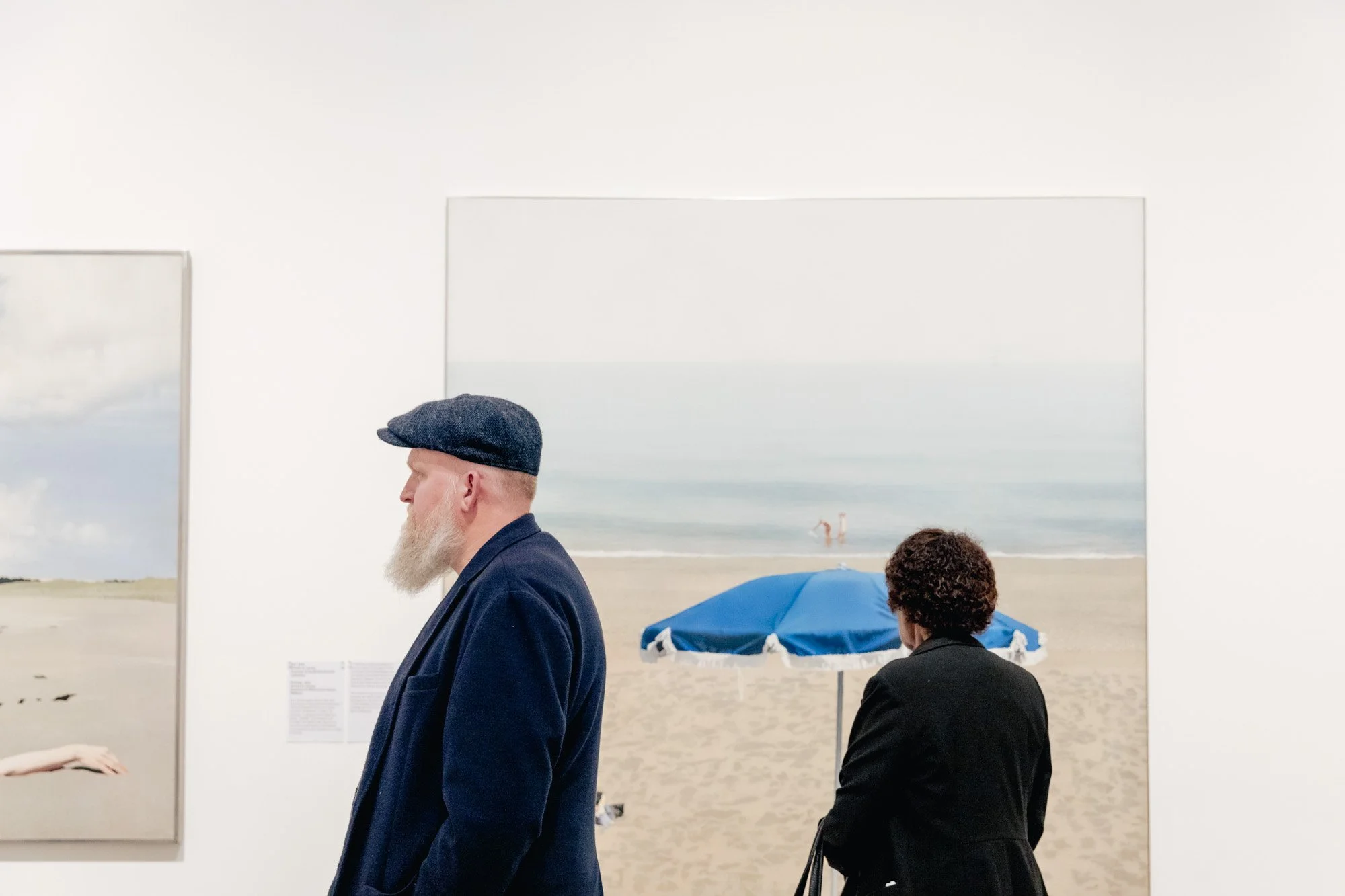 Two people looking at beach-themed artwork in an art gallery. One man with a grey beard, wearing a dark coat and a flat cap, is in the foreground. The other person, with curly hair and a dark coat, is in the background, facing away. The background ar