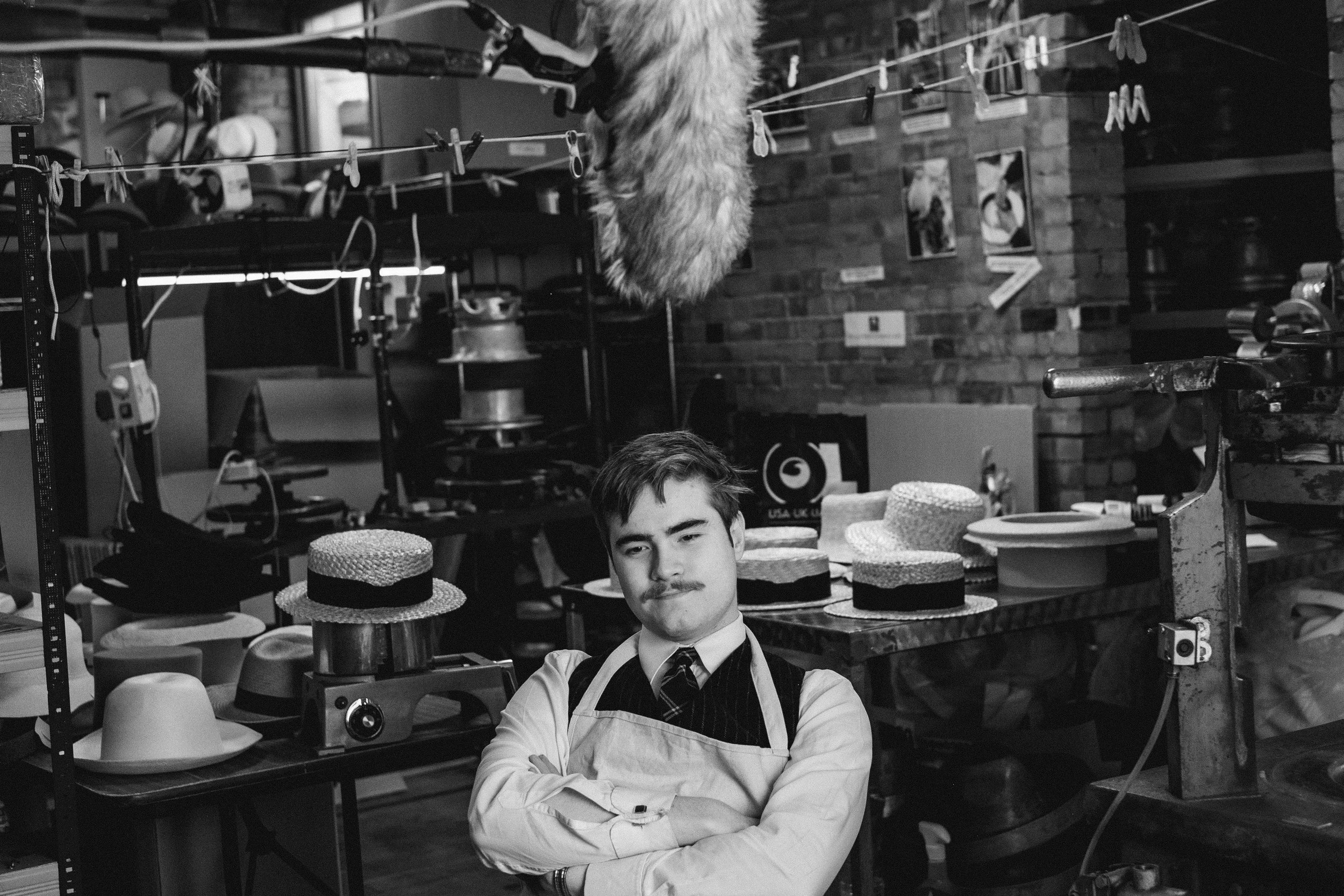 A man standing with arms crossed in a hat shop, surrounded by hats, sewing equipment, and a rustic interior.