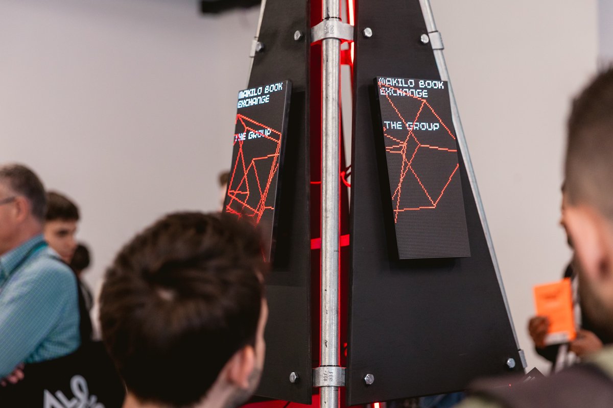 Two electronic screens showing a game of chess with red lines, mounted on a black and silver stand, with people watching in the background.