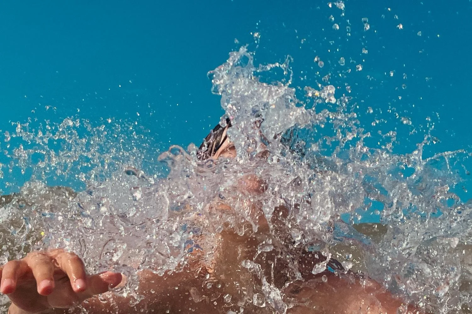 A person swimming underwater in a pool, with splashing water surrounding them and another person's hand visible in the foreground.