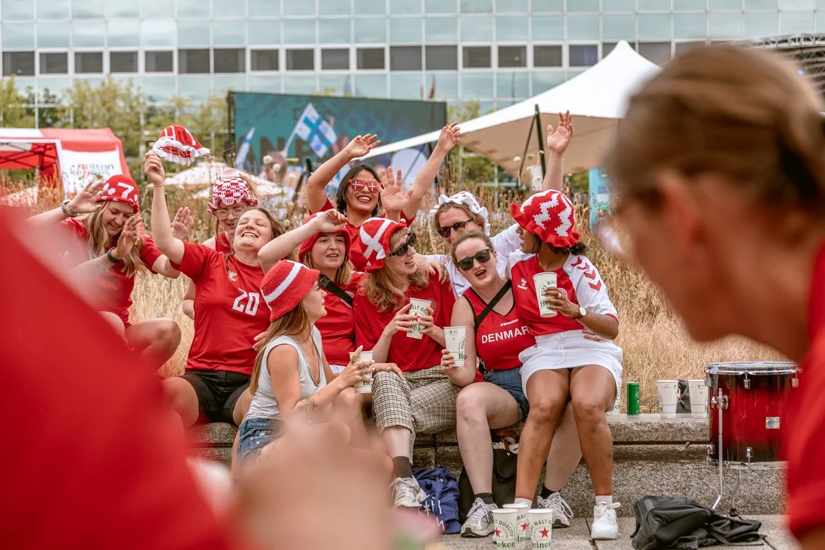Group of people sitting outdoors in Denmark fan attire, cheering and enjoying drinks, with some wearing red and white hats, in front of a building with glass windows and a large screen.