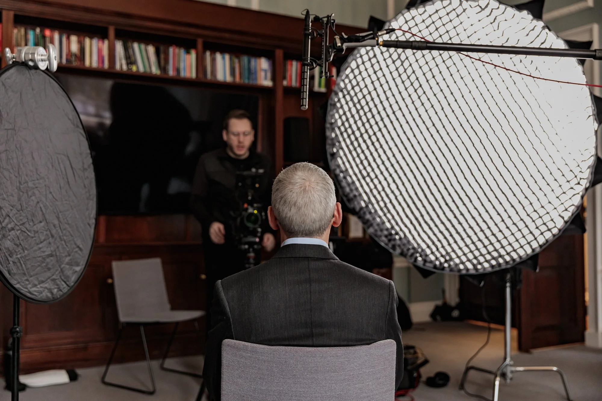 An older man with gray hair wearing a dark suit sits in front of a camera recording a video interview or speech. A videographer in black is behind the camera, and large studio lights are set up on either side.
