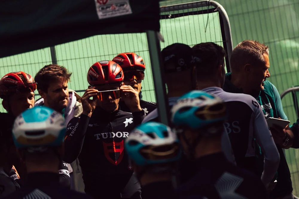 Cyclists and team staff behind a green fence at a cycling event, with some wearing red helmets and team gear.