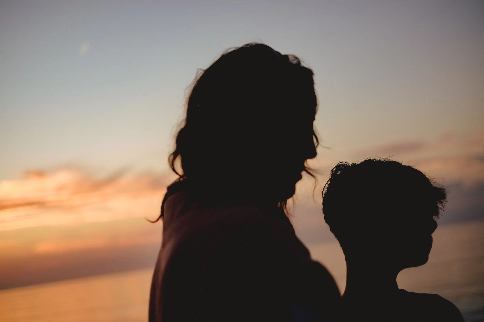Silhouettes of two people, one with long hair and one with short hair, facing each other at sunset near the water.